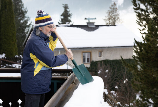 An adult gray-haired man stands on the balcony and cleans snow from the railing with a shovel