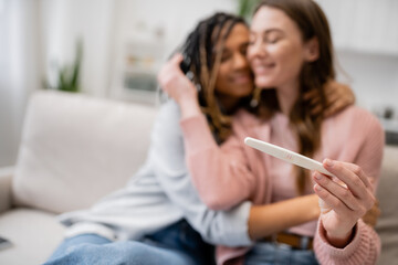 Fototapeta premium cheerful african american and lesbian woman hugging girlfriend with pregnancy test on blurred background.