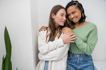 joyful interracial and lesbian women smiling while hugging near green plant at home.