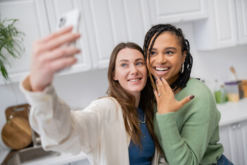 lesbian woman taking selfie while happy african american girlfriend showing engagement ring on finger.