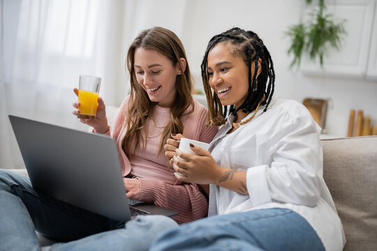 young african american lesbian woman holding cup of coffee near happy girlfriend using laptop while working from home.