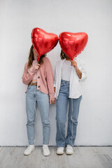 interracial lesbian women covering faces with red heart-shaped balloons on grey.