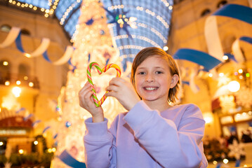 happy child holding heart shaped candy stands in front of christmas decorated tree in winter at mall in evening