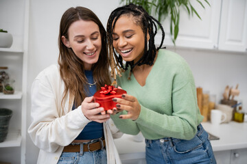 happy interracial and lesbian women holding red heart-shaped gift box with bow on valentines day.