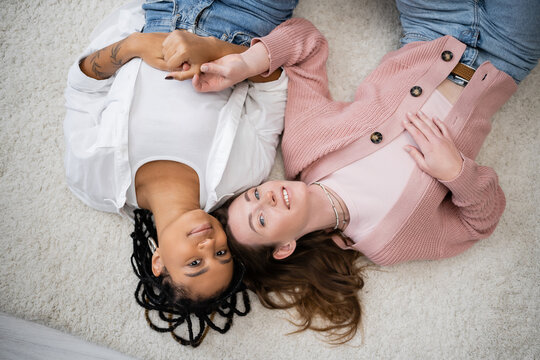 top view of joyful and multiethnic lesbian couple lying on soft carpet.