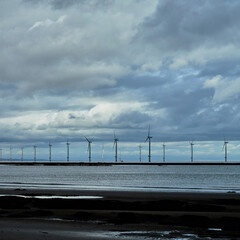 Cloud shadows play across a near-shore wind farm in North-East England, huge cumulus formations driven by the strong wind turning the massive turbines.