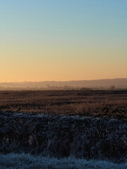 Dawn sunlight casts long shadows across a muted, frost and mist shrouded winter landscape of hills, trees, fields and huge sky.