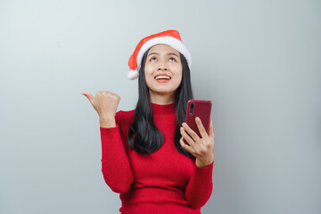 Pretty lovely lady asian thai women in red sweater and santa christmas hat isolated over grey background. Holding and using mobile smartphone or cellphone