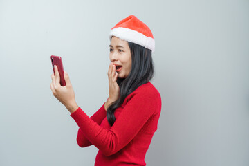 Pretty lovely lady asian thai women in red sweater and santa christmas hat isolated over grey background. Holding and using mobile smartphone or cellphone