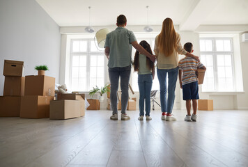 Fototapeta na wymiar Happy family with children on moving day standing between cardboard boxes inspecting their new home. Rear view of young family enjoying their new apartment. Mortgage, relocation and moving day concept