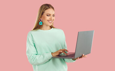 Happy blondy woman in mint sweatshirt doing remote job working on laptop on pink background and looking at screen. Distance work, online shopping, browsing social media, internet surfing concept.