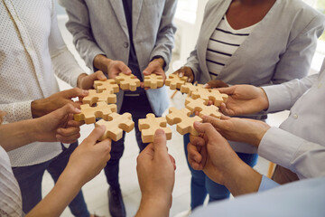 Group of men and women standing in circle and connecting identical wooden puzzle pieces. Jigsaw puzzle pieces symbolizing teamwork in hands of unrecognizable multiracial people. Cropped image.