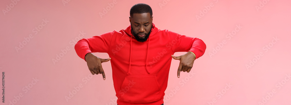 Wall mural Focused interested serious-looking young african american bearded strong guy wearing red hoodie bend head look pointing down curiously, intrigued what happening lower floor, standing pink background