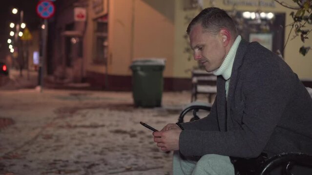 Young handsome man smiles as he sends text sms message using his smartphone sitting on bench at snowy winter night. Posting social media, online shopping, texting, e commerce