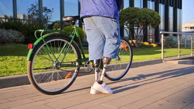 Slow motion view of a man with prosthetic legs. Walking with a bicycle on the street with greenery