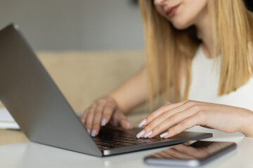 Cropped view of blurred coach using laptop near mobile phone with blank screen at home.