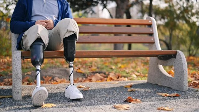Slow motion view of a man with prosthetic legs and white sneakers. Sitting on a bench in a park. Fallen yellow leaves around