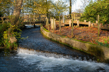 Fototapeta premium water cascading over a small weir in the river with an old wooden bridge in the background