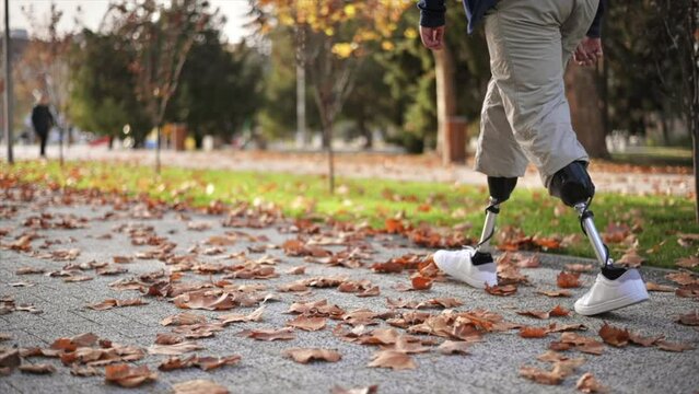 Slow motion view of a walking man with prosthetic legs and white sneakers in a park. Fallen yellow leaves on the ground