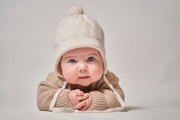 A portrait of an infant in knitted clothes and a white cap looks at the camera