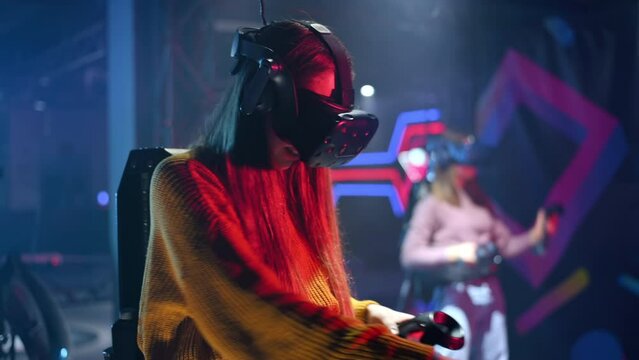 A joyful, interested girl is playing on VR treadmill with helmet, controllers and a backpack. Other people playing nearby