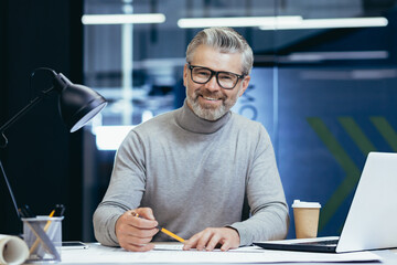 Portrait of an older gray-haired male teacher. He sits at a desk with a laptop, looks at the camera, smiles, holds a pencil in his hands. Teaches online, remotely.
