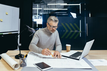 General photo of the workplace of a gray-haired senior man, a designer, architect, engineer. He sits at the desk in the office. He draws a project, a plan with a pencil and a ruler on a roll of paper.