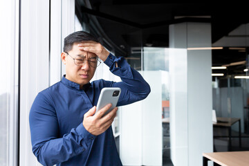 Upset asian man in office at work reading bad news from phone online, businessman in shirt near window working inside office, using smartphone.