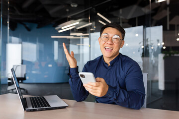 Portrait of happy Asian businessman, man celebrating achievement good results and victory, boss looking at camera inside office holding phone, entrepreneur working inside office.
