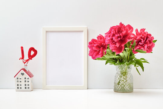 Mockup with a white frame and red peonies in a vase, red heart and white house on a white table.  Valentines Day, Happy Women's Day concept