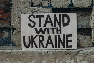 Ukrainian protest against war with banner placard  with inscription message text Stand With Ukraine, ruined city background. Crisis, peace, Russian aggression invasion concept. anti-war demonstration.