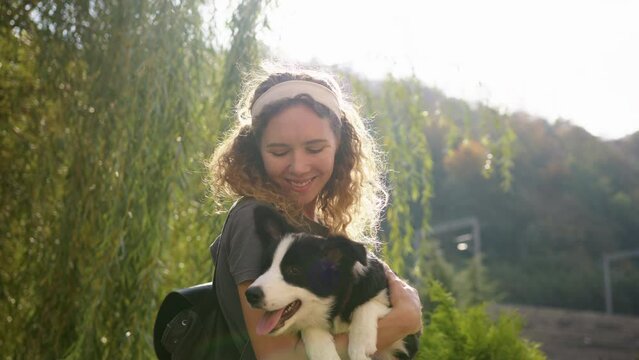 Girl with a border collie puppy in her arms kisses him and hugs him by a tree in the sunlight against the background of mountains