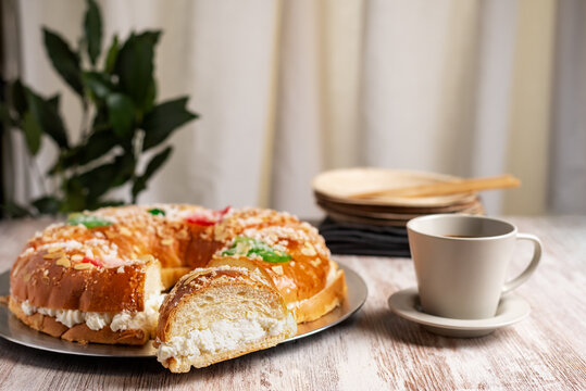 Roscon de reyes with a cut piece revealing the cream of its filling, along with a cup of coffee, in the background several wooden serving dishes.