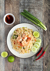 Thai fried rice with shrimp, green onion, lime, chili and fish sauce in a white plate on a wooden table, top view.