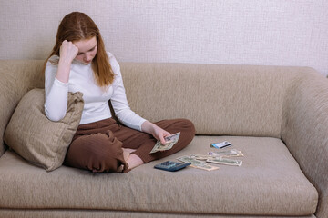 A young upset girl is counting the money of her budget sitting at home on the couch