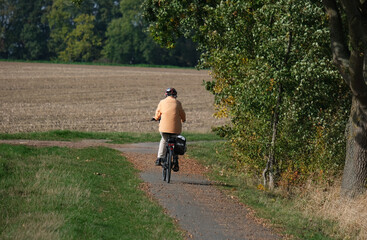 Ältere Frau mit Helm, Jacke und Satteltasche fährt auf einem Fahrrad auf einem Radweg in der Natur an Wäldern und Feldern vorbei