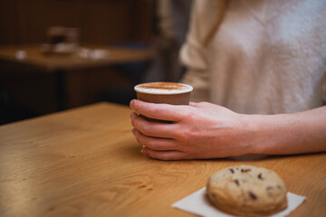 Close up shot of cup of coffee held by young woman and a chocolate cookie inside a coffee shop
