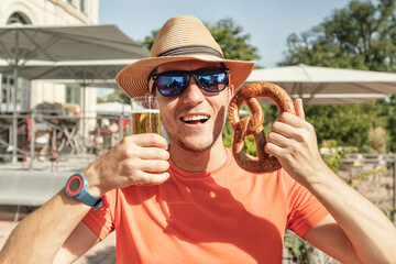 Happy tourist man drinking glass of beer and biting a traditional German snack pretzel in...