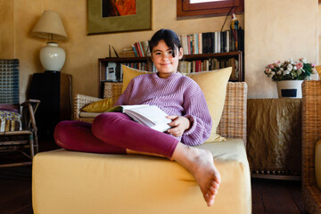 similing girl in lilac pullover reading a book in yellow armchair
