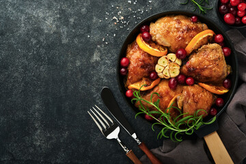 Baked chicken thighs with orange, cranberry and spicy herbs rosemary servered in frying pan on black concrete background. Festive Christmas Dinner Concept menu. Top view. copy space