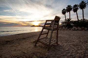 Silla del socorrista en la playa de Benidorm con el sol al atardecer de fondo mirando al horizonte del mar Mediterráneo con las palmeras.
