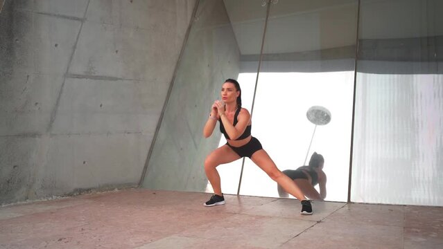 Fit middle age woman doing side lunges exercising outdoors. Female athlete warming up muscles and stretching while standing near mirror wall. Confidence sporty female training alone