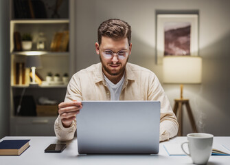 Young caucasian male in eyewear working on laptop at home, remote employment.