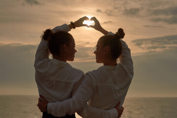 sun in shape of heart from hands of twins sisters on morning sea and sky background