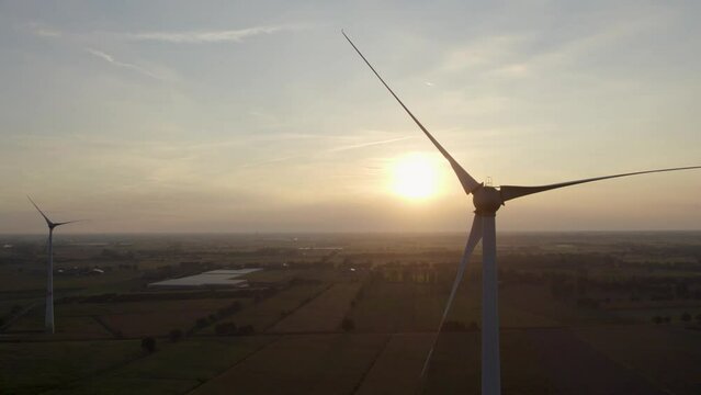 Aerial shot by a drone of windmills with rotating wings among green fields against a sunrise or sunset background. Wind farm with turbine cables for wind energy. Renewable energy source, earth care