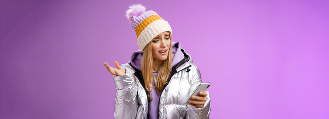 Obraz na płótnie Canvas Upset disappointed attractive whining gloomy blond girl in silver jacket standing outside hat holding smartphone shrugging raising hand dismay complaining slow mobile internet, purple background