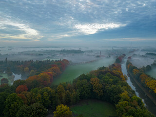 Aerial view of foggy trees in field at colorful sunrise in autumn. Colorful landscape with forest in low clouds, river, meadow in fog, orange sky with sun in the morning in fall. Top view. Nature