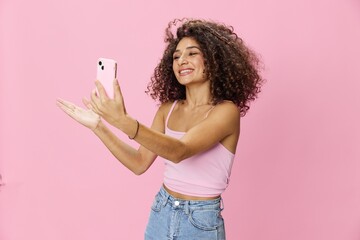 Woman blogger holding phone in hand video call, with curly hair in pink top and jeans poses on pink background, copy space, technology and social media, online