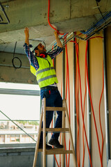 An experienced electrician who stands on a ladder and adjusts cable bellows for the electrical network of the building where he works.