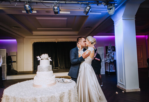 Stylish groom and beautiful blonde model bride kiss in a restaurant near a sweet cake in a restaurant. Wedding photography of newlyweds, food.